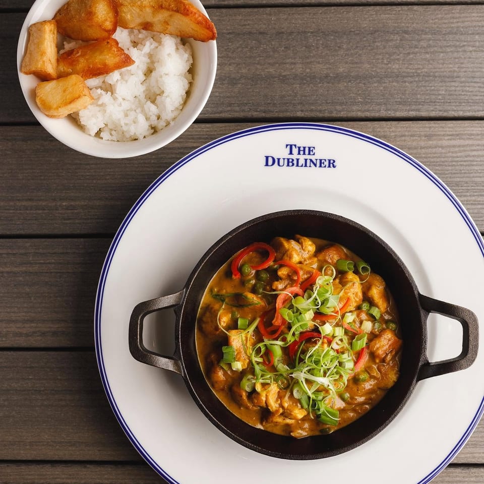 Top-down view of a spicy garnished stew in a handled skillet beside white rice and fried plantains on The Dubliner plate.