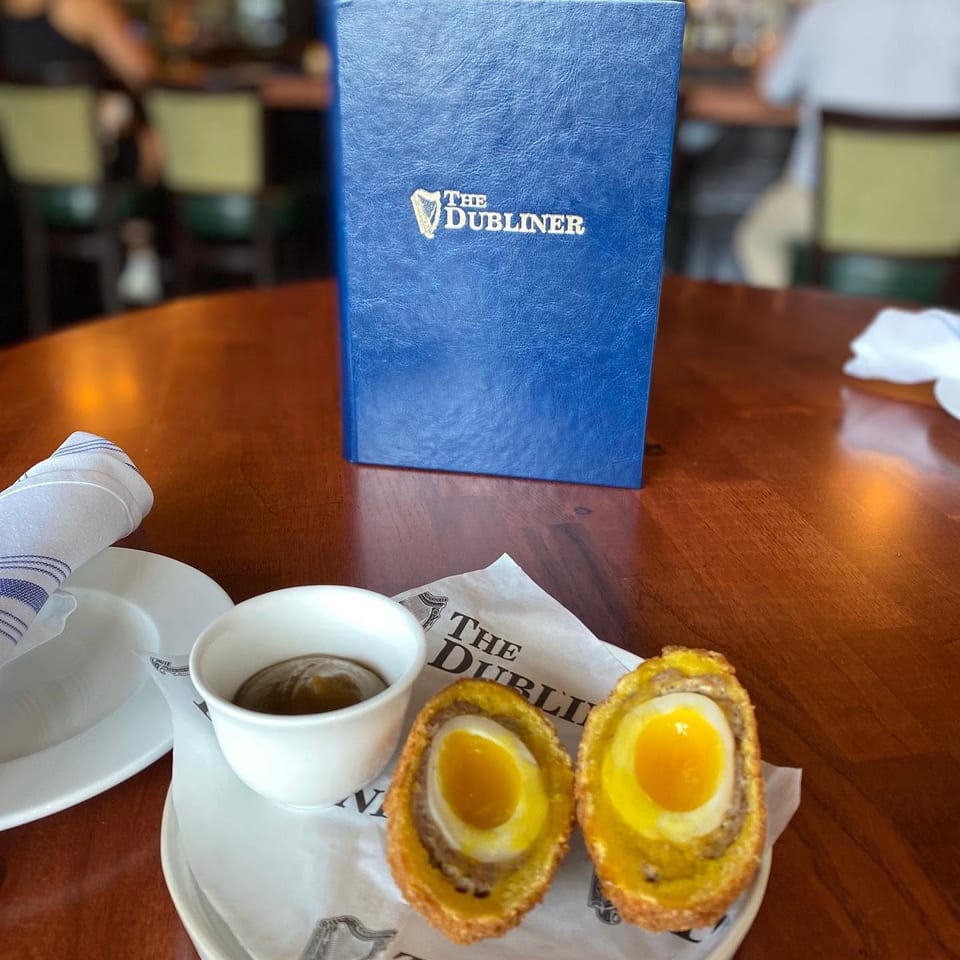 Blue The Dubliner menu beside two halved, breaded Scotch eggs with dipping sauce on a restaurant table.