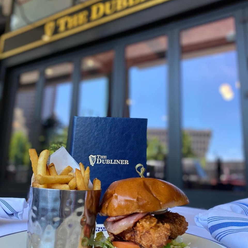 The Dubliner restaurant exterior behind a plated fried chicken sandwich with bacon, lettuce, tomato, and fries.