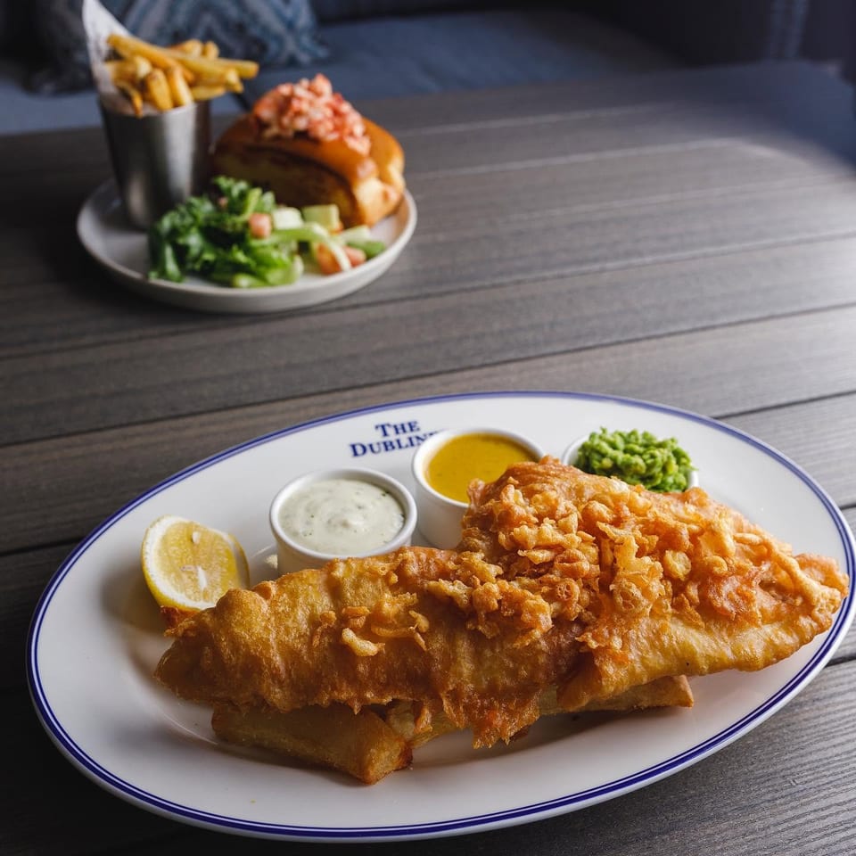 Battered fish and chips with lemon, tartar sauce, and mushy peas, accompanied by a burger, fries, and salad.