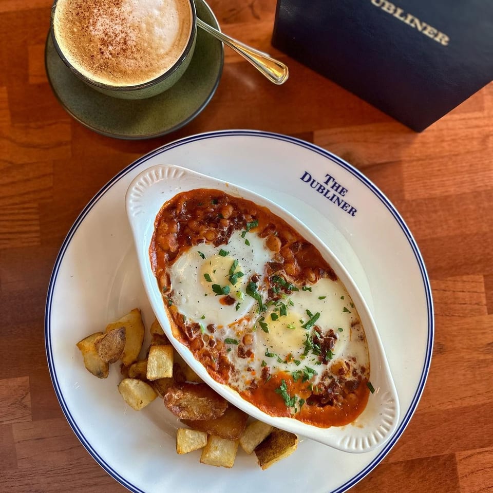 The Dubliner plate of cheesy baked beans with potatoes, accompanied by a frothy cappuccino on a wooden table.