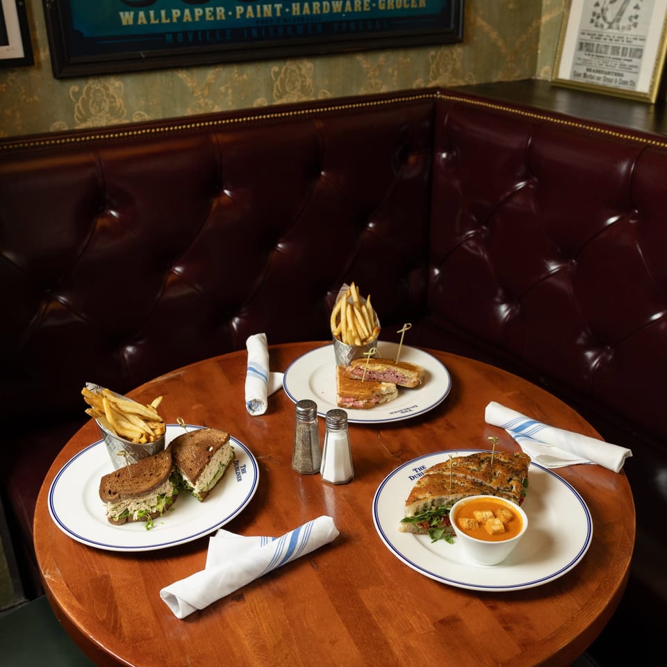 Restaurant booth with three plated sandwiches, fries, dipping sauce, rolled napkins, and vintage Cooke’s wall signage.